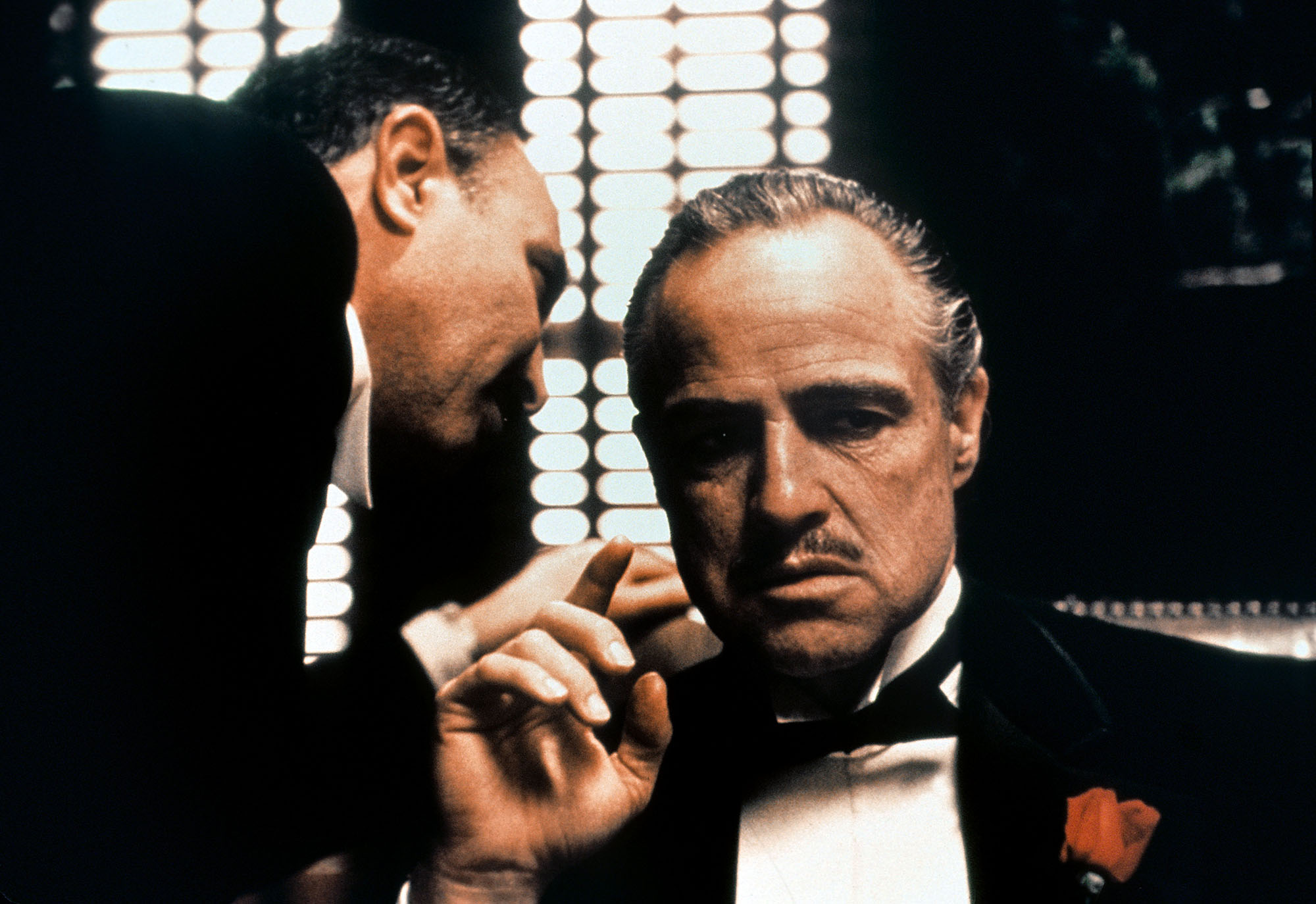 image from the 1972 movie "The Godfather." On the left is Salvatore Corsitto as Amerigo Bonasera. He is whispering into the ear of Vito Corleona (Marlon Brando, seated at a desk and wearing a tuxedo).