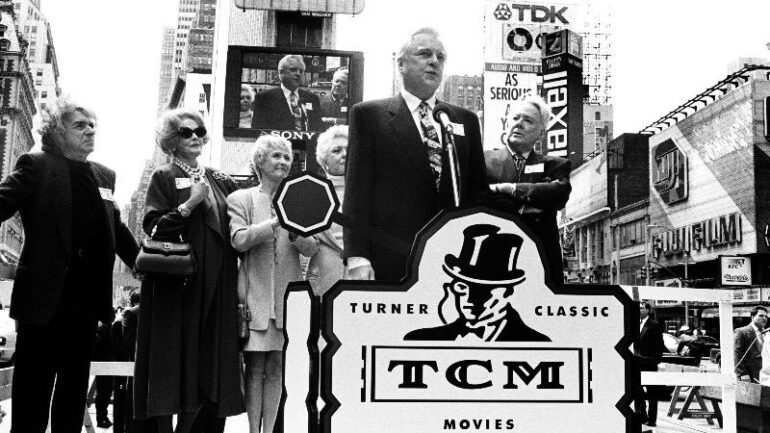 a black-and-white 1994 photo of Robert Osborne standing at a lectern and speaking during the launch announcement of Turner Classic Movies in April 1994.