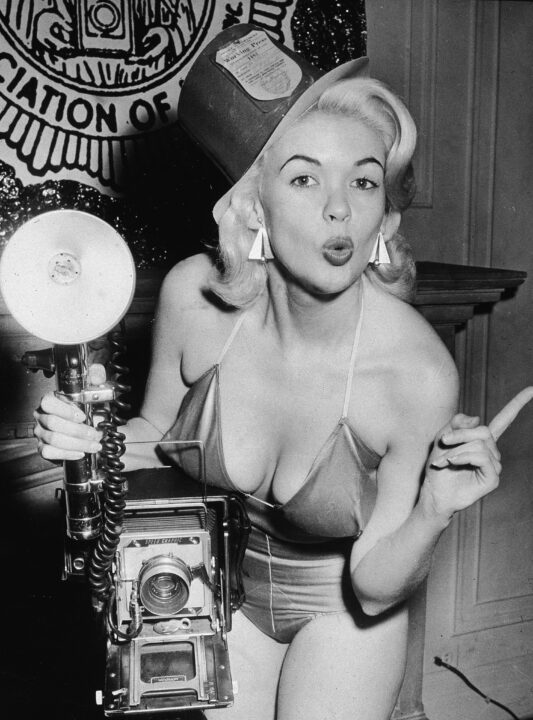 American film actor and sex symbol Jayne Mansfield (1933 - 1967) purses her lips and holds a camera while dressed in a bathing suit and a paper hat labelled 'Working Press,' 1956. 