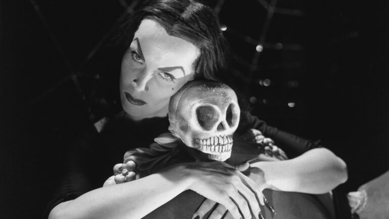 Promotional portrait of Finnish-born American actress Maila Nurmi (born Maila Syrjaniemi, 1922 - 2008), in character as Vampiria, as she poses with her arms around the skull-topped back of a chair, 1950s. Nurmi developed the Vampira character when she was hired to host television braodcasts of horror movies on the Los Angeles station KABC-TV; she later played the character in the film Ed Wood-directed film, 'Plan Nine from Outer Space.'