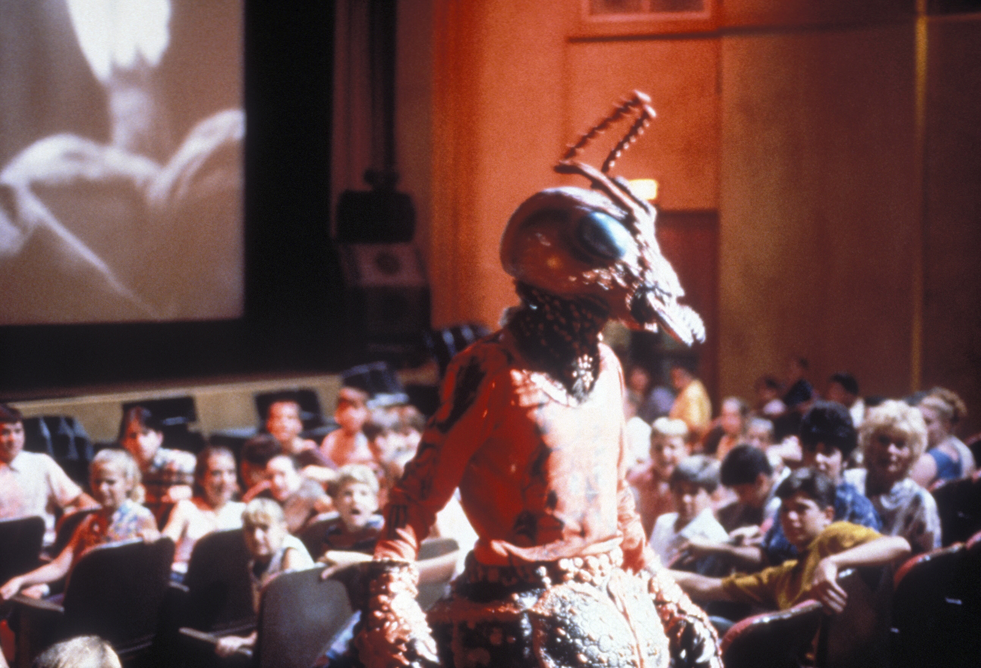 image from the 1993 movie "Matinee." It is a scene in an early '60s movie theater, where a bunch of children are watching a horror movie called "Mant!" Running in the aisle of the theater, trying to scare the kids, is someone dressed as an ant-monster.