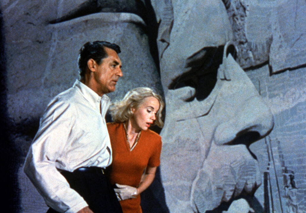 image from the 1959 movie "North by Northwest." On the far left of the horizontal image is star Cary Grant, wearing a white dress shirt and black trousers. His left arm is around costar Eva Marie Saint, wearing a red dress, and he is holding her close as both look down with concern from where they are standing atop the Mount Rushmore monument in the film's climax. Behind Saint and a little to her left, the carved-stone face of Teddy Roosevelt on the monument can be seen.