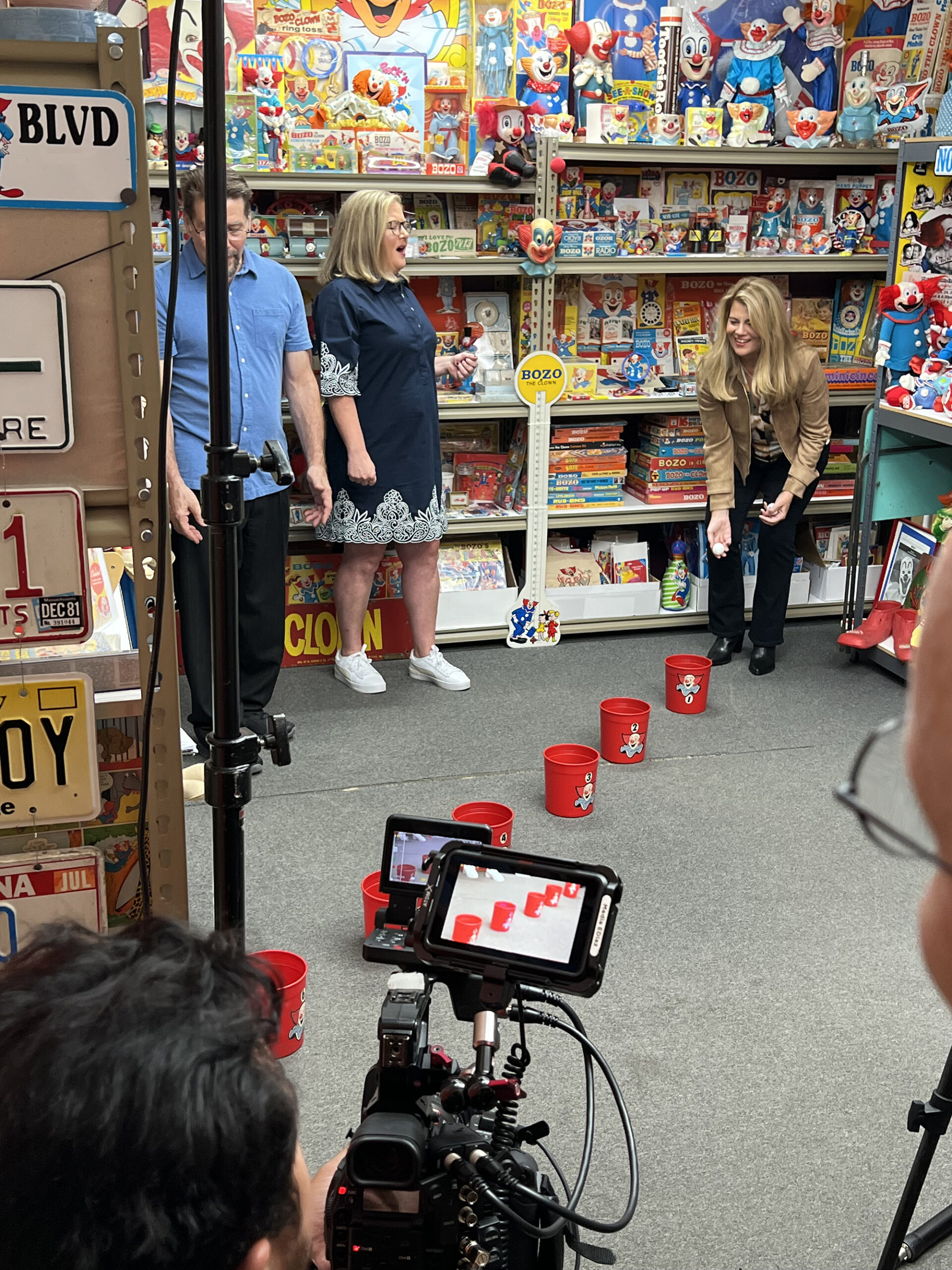 image from a Bozo the Clown-themed episode of MeTV's "Collector's Call." Host Lisa Whelchel is playing Bozo's famous "Grand Prize Game" (throwing ping pong balls into increasingly farther away buckets) as appraiser Kate Martin and Bozo collector Tommy Holbrook look on.
