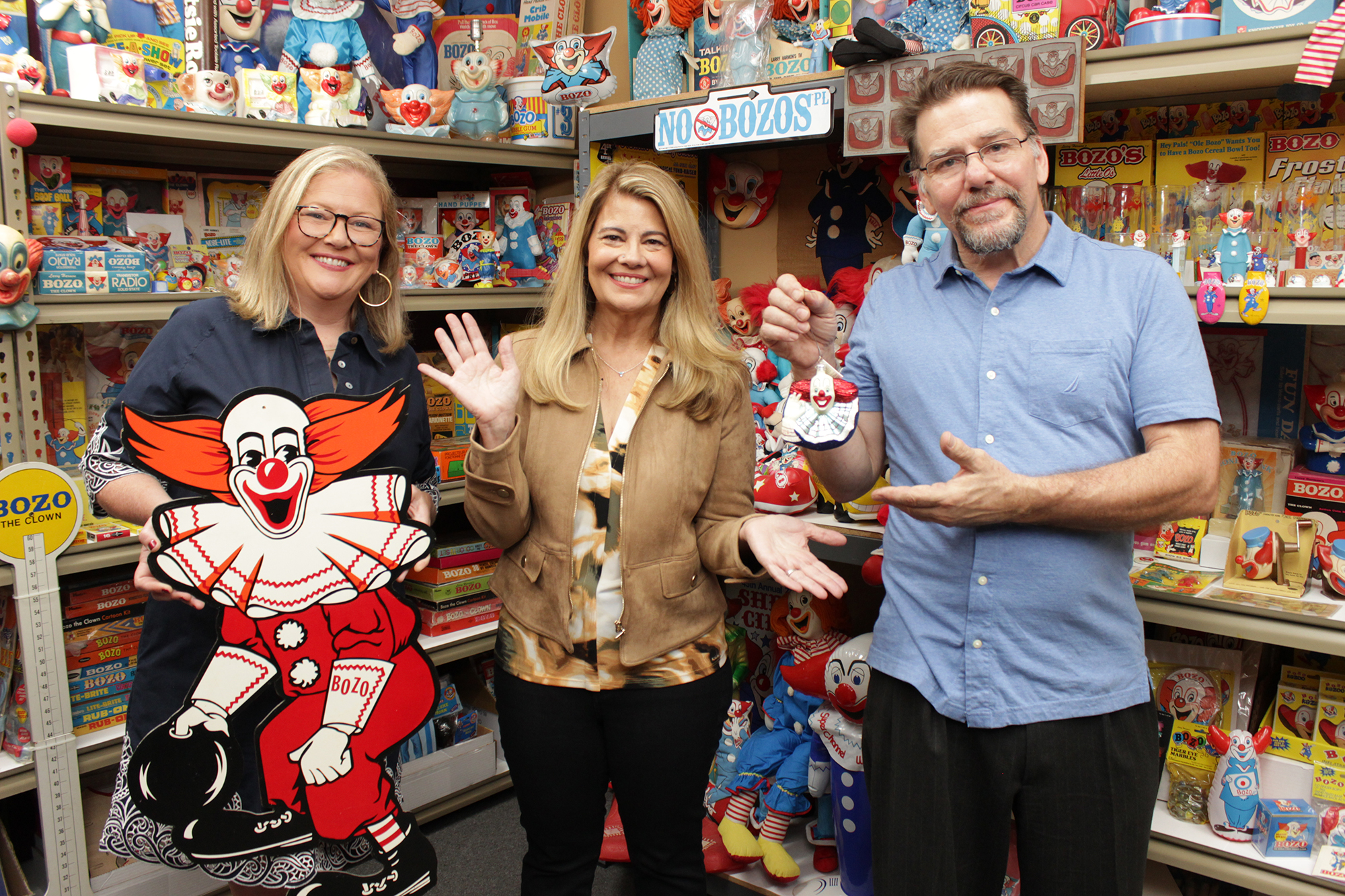 image from a Bozo the Clown-themed episode of MeTV's "Collector's Call." From left to right, each holding Bozo memorabilia, are appraiser Kate Martin, series host Lisa Whelchel and collector Tommy Holbrook