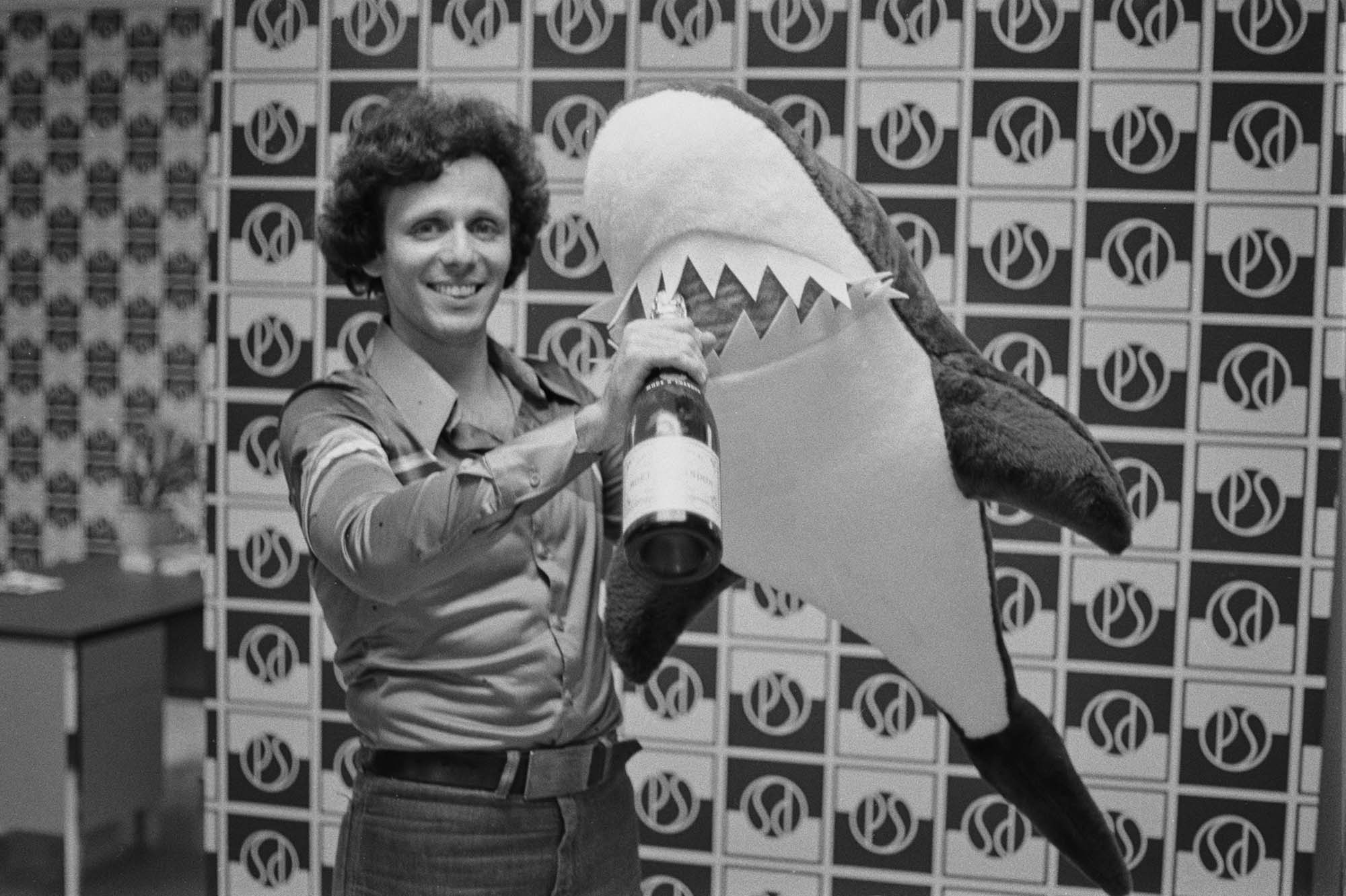 black-and-white archival 1975 photo of music producer Dickie Goodman, standing and holding a bottle of champagne (in his right hand) up to the mouth of a stuffed toy shark in his left hand and smiling in a promo shot for the success of his novelty song 