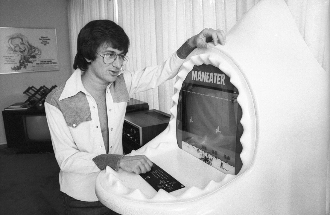 black-and-white photo of director Steven Spielberg posing for a photo with a 'Maneater' arcade video game cabinet at his Universal studios office in December, 1975 in Los Angeles, California. Spielberg is wearing a collared shirt that is unbuttoned at least down to his navel and wearing glasses as he rests both arms on the cabinet and looks at the video game screen with a bit of amusement.