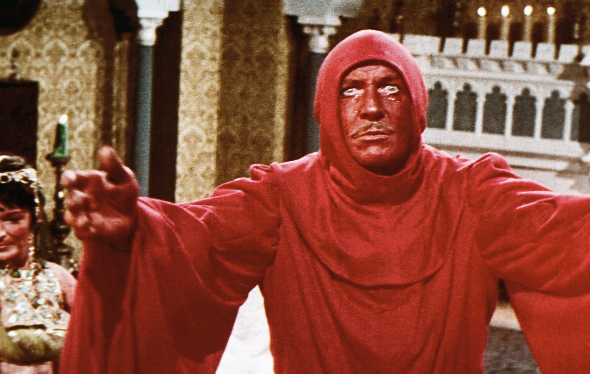 scene from the 1964 movie "The Masque of the Red Death." Vincent Price, as Prince Prospero, is reaching out with open arms toward the camera, with an intense and fearful look in his eyes. He is is clad in an all-red robe and hood, and his face is painted red.