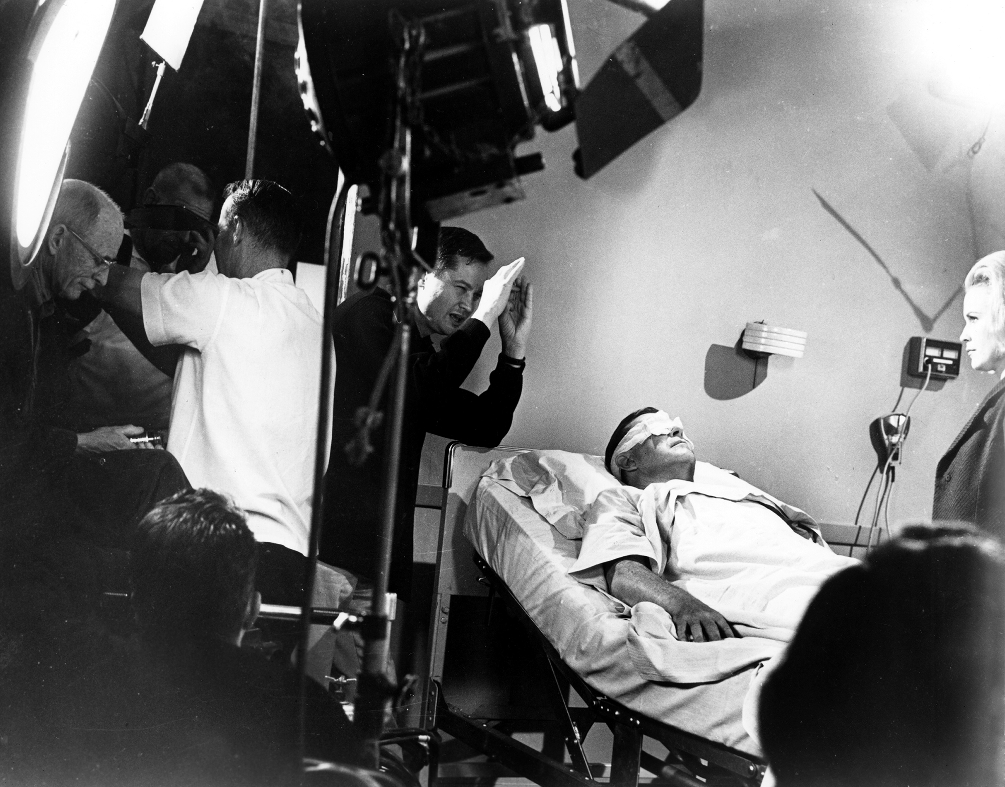 black and white behind-the-scenes image from the 1963 movie "X: The Man With the X-Ray Eyes." Director Roger Corman is standing on the left with his hands up to his face to envision the blocking for a scene from the film. Lying just below him in a hospital bed is star Ray Milland, with his eyes covered with bandages.