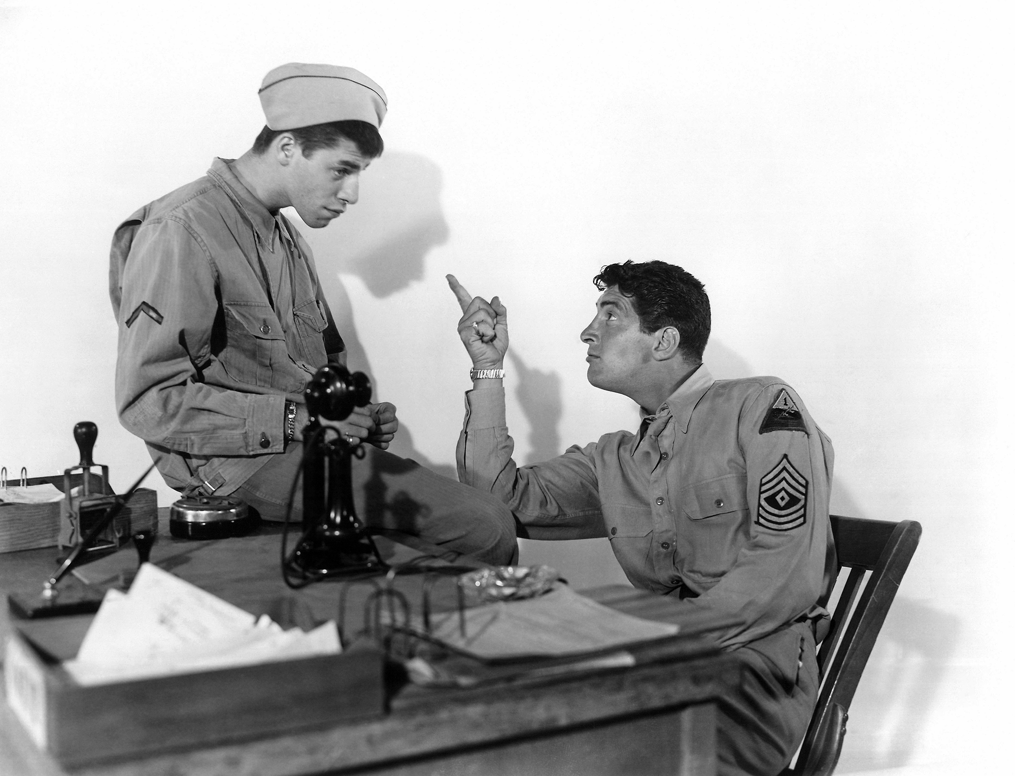 black and white photo from the 1950 movie "At War With the Army." Star Dean Martin, dressed in a superior officer military uniform, is seated at a desk, looking up and pointing angrily at costar Jerry Lewis, dressed in a private's uniform and sitting on the desk with a confused look on his face.