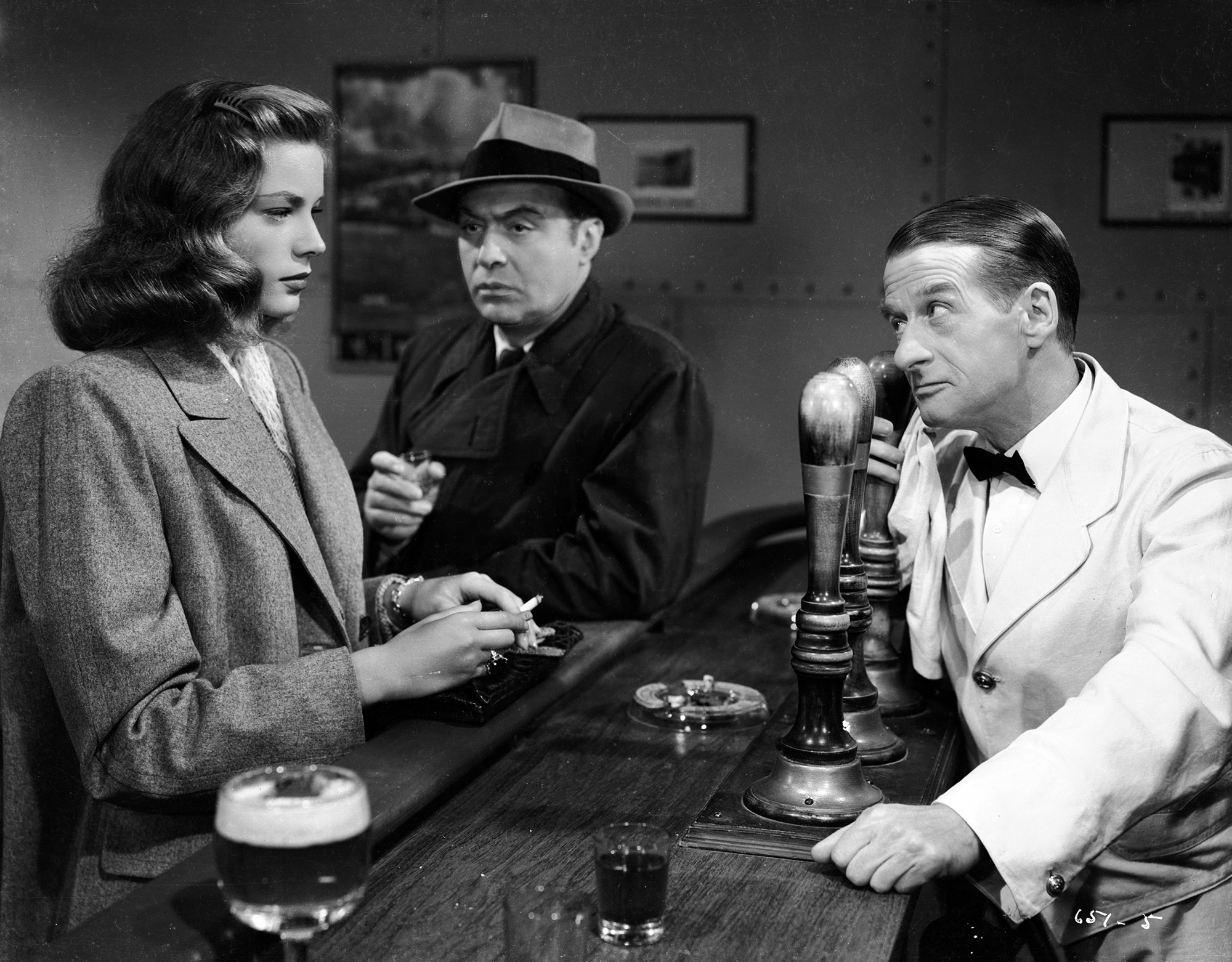 Medium black and white shot from the 1945 movie "Confidential Agent" of Lauren Bacall as Rose Cullen with coat holdingcigarette standing at bar across from Alec Harford as Bartender with Charles Boyer as Luis Denard with coat and hat standing holding drink leaning on the bar between them.