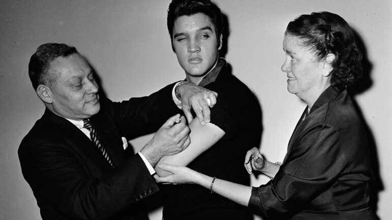 Backstage at the 'The Ed Sullivan Show,' American singer and musician Elvis Presley (1935 - 1977) (center) glances out of the corner of his eye at a smiling nurse while a doctor injects him with a polio vaccine, New York, New York, October 28, 1956. (Photo by CBS Photo Archive/Getty Images)