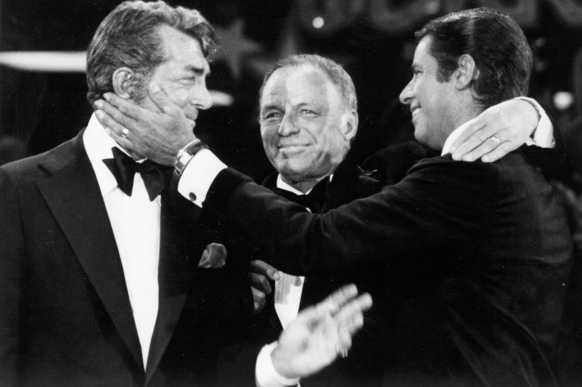 (L to R) Dean Martin, Frank Sinatra and Jerry Lewis perform during the 1976 telecast of The Jerry Lewis MDA Telethon in Los Angeles, California. They are all dressed in tuxedos, and Lewis is reaching out and touching Martin's right cheek as Martin reaches back. They all look emotional and happy.