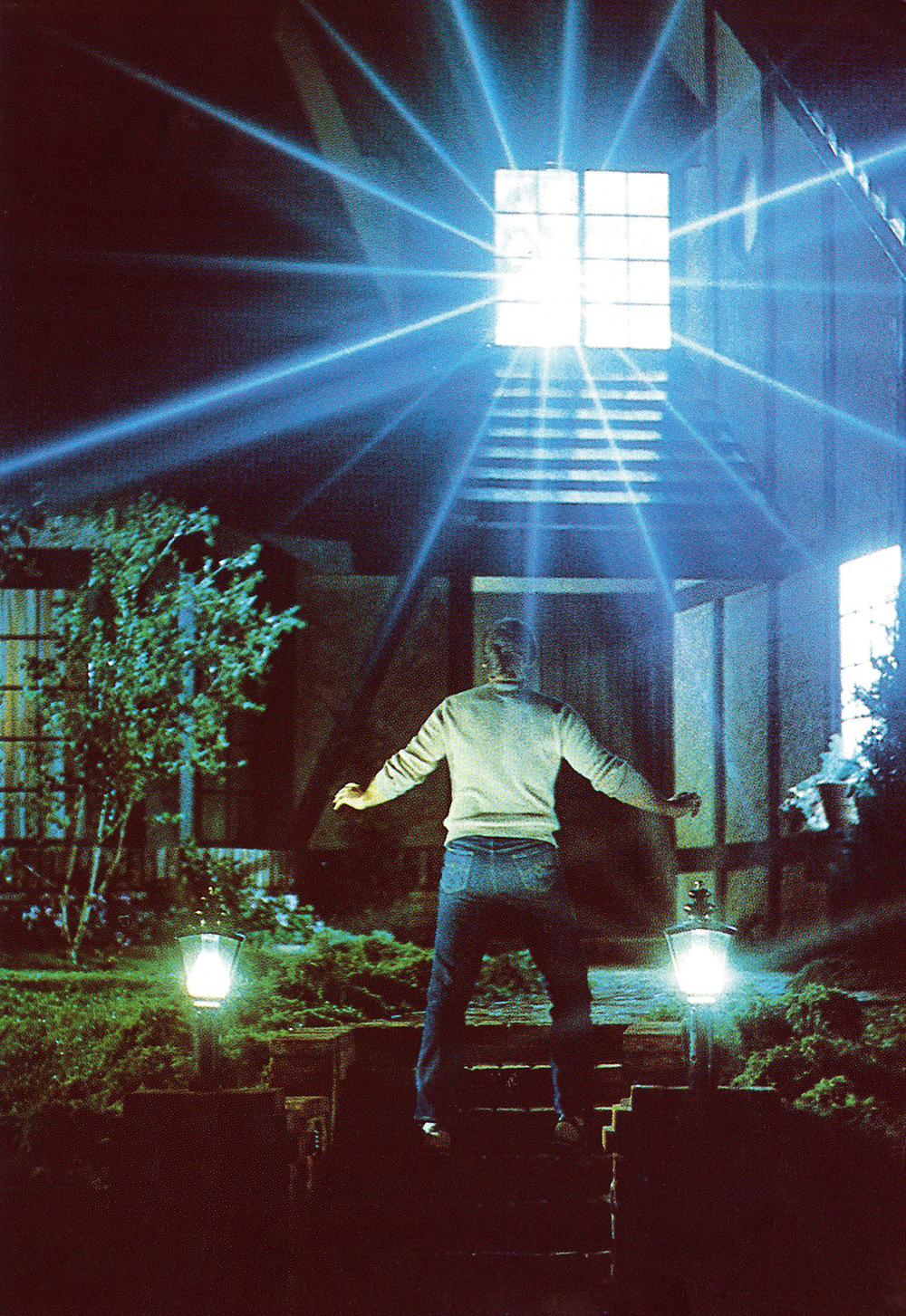 image from the 1982 movie "Poltergeist." It it a night scene, with star Craig T. Nelson being seen from the back as he climbs the front outside steps at the front of the house. He is halted in alarm as a bright flashing blue light is erupting from a window just above the front door.