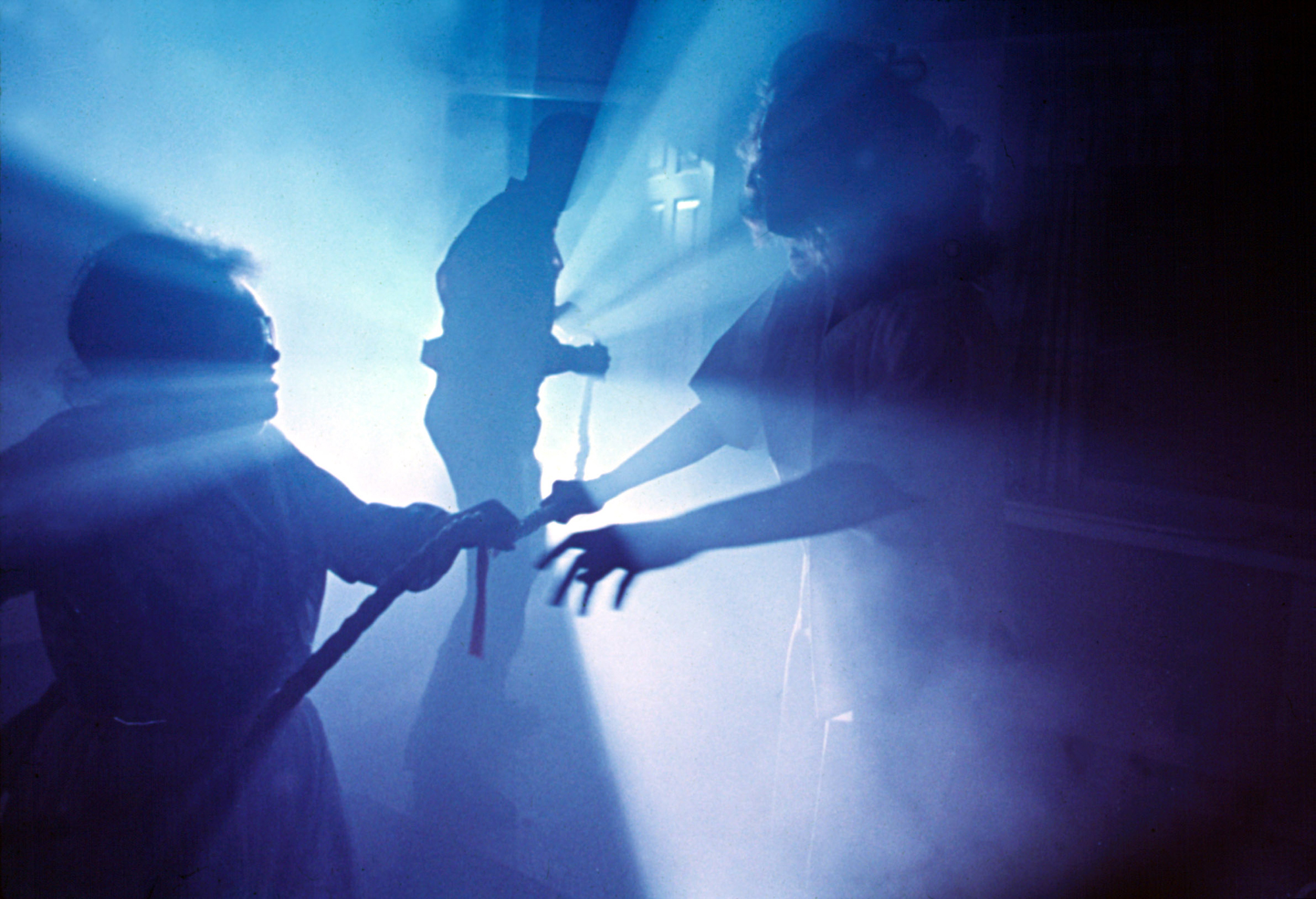 image from the 1982 movie "Poltergeist." It is a shot in a bedroom with an open closet door, out of which is emanating a bright light bathing the entire room in blue. The shadow of star Craig T. Nelson can be seen in the background, holding a rope leading into the closet. Shadows of costars Zelda Rubinstein (on left) and JoBeth Williams (right) are seen in the foreground.