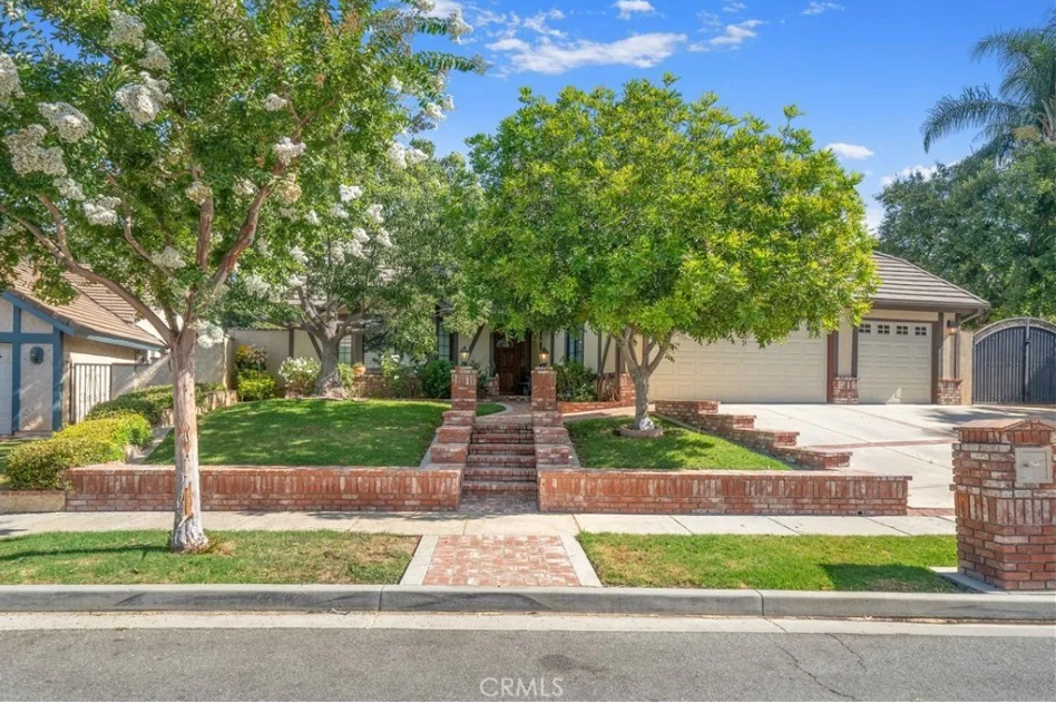 wide shot photo of the front exterior of the real home used during the filming of the 1982 movie "Poltergeist." A brick walkway leads up to the house, which has several flowering trees in front somewhat obscuring the front windows. The home's three-car attached garage can be seen on the photo's right.