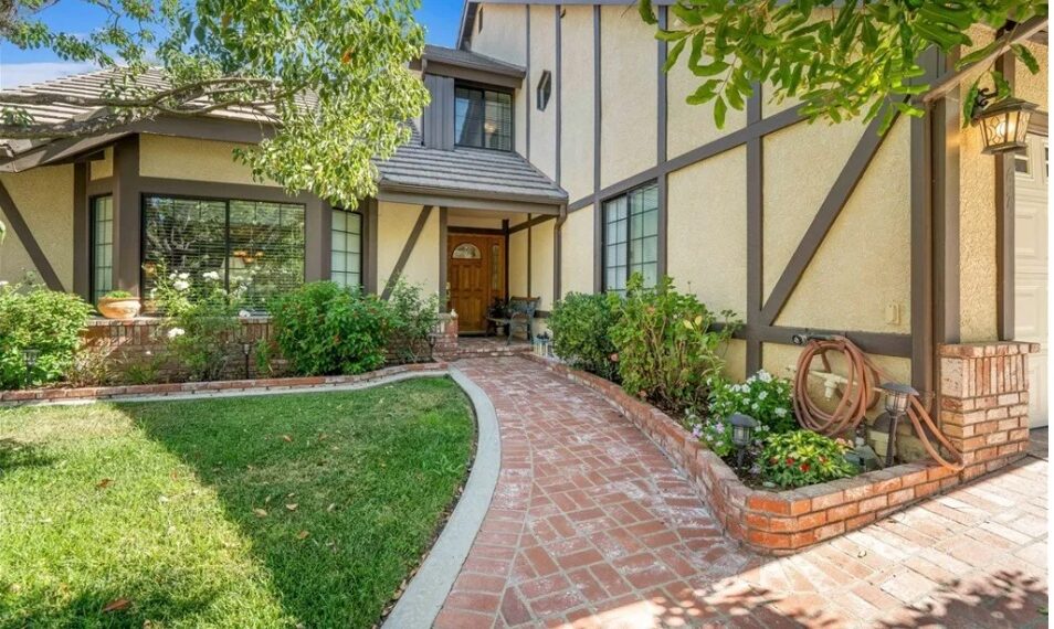 image of the front exterior of the house in California where much of the movie 'Poltergeist' was filmed, which is now up for sale. It's a traditional house style, with wood and stucco, and a brick walkway leading up to the front door. Nicely trimmed trees and bushes decorate the front yard.