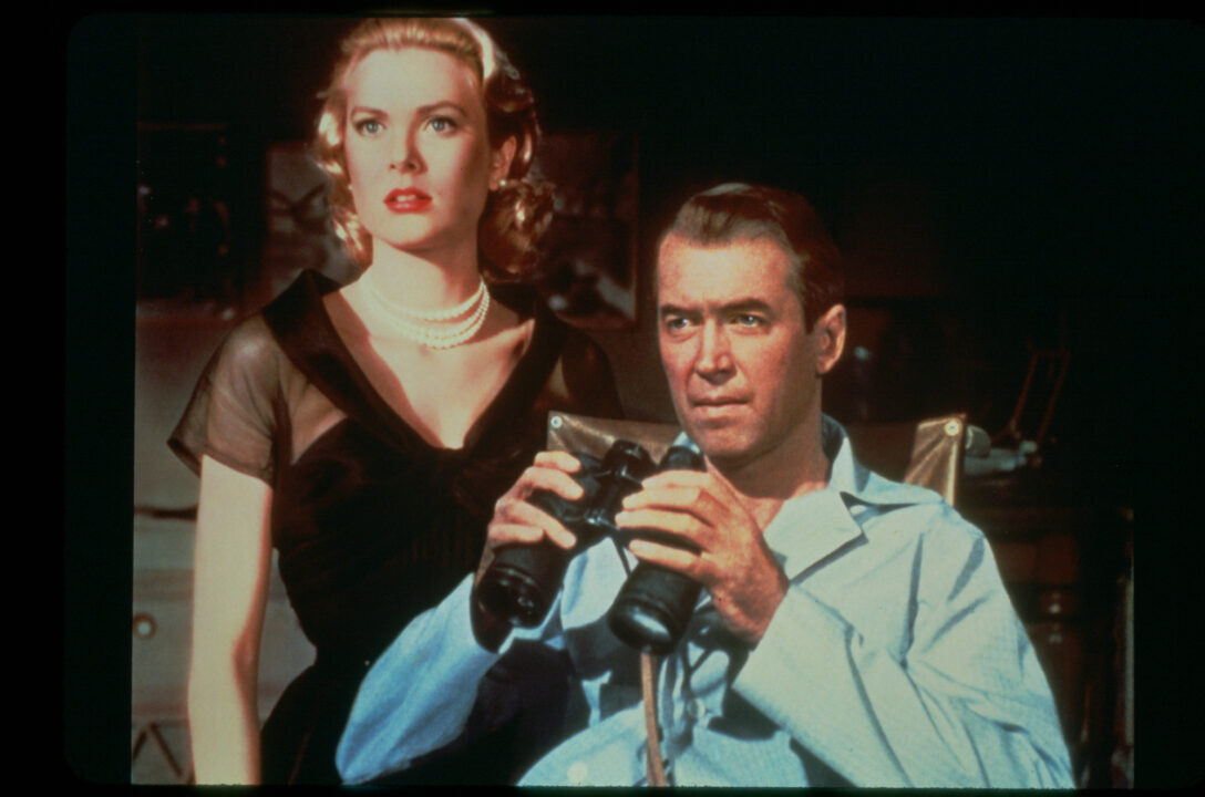 image from the 1954 movie "Rear Window." It is a medium shot of stars Grace Kelly and James Stewart. Kelly is standing on the left, wearing a black, lower-cut dress. Right next to her, on her left (right in the photo), seated in a wheelchair is Stewart, in blue pajamas and holding binoculars in both hands. They are both looking with concern at something to their right (left in photo) that cannot be seen by us.