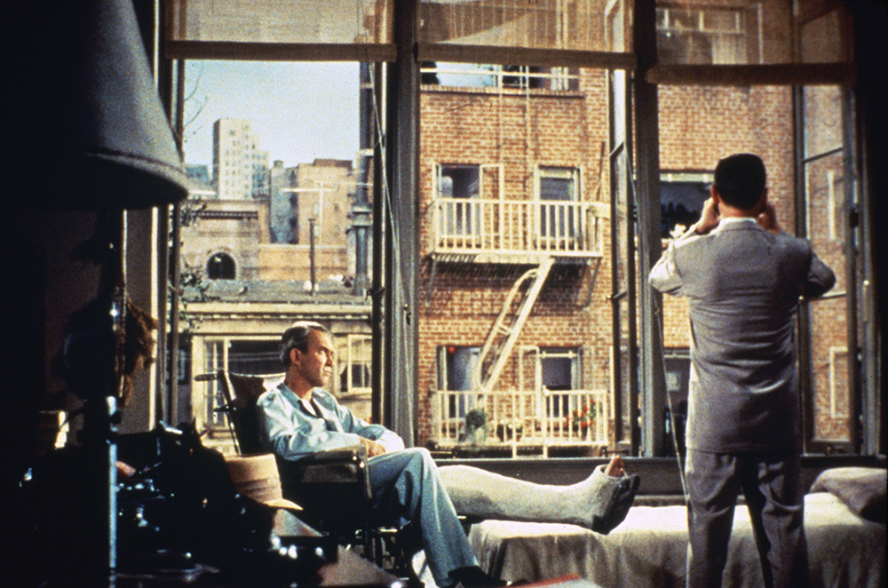 image from the 1954 movie "Rear Window." The scene is an a NYC apartment with a wide window looking out on a courtyard, across which is another apartment building with windows. Seated on the left is star James Stewart, in a wheelchair with his left leg in a cast and outstretched on a rest. He is looking serious as costar Wendell Corey, on the right and with his back to us, wearing a suit and fedora hat, is holding binoculars to his eyes and looking at the windows across the way.