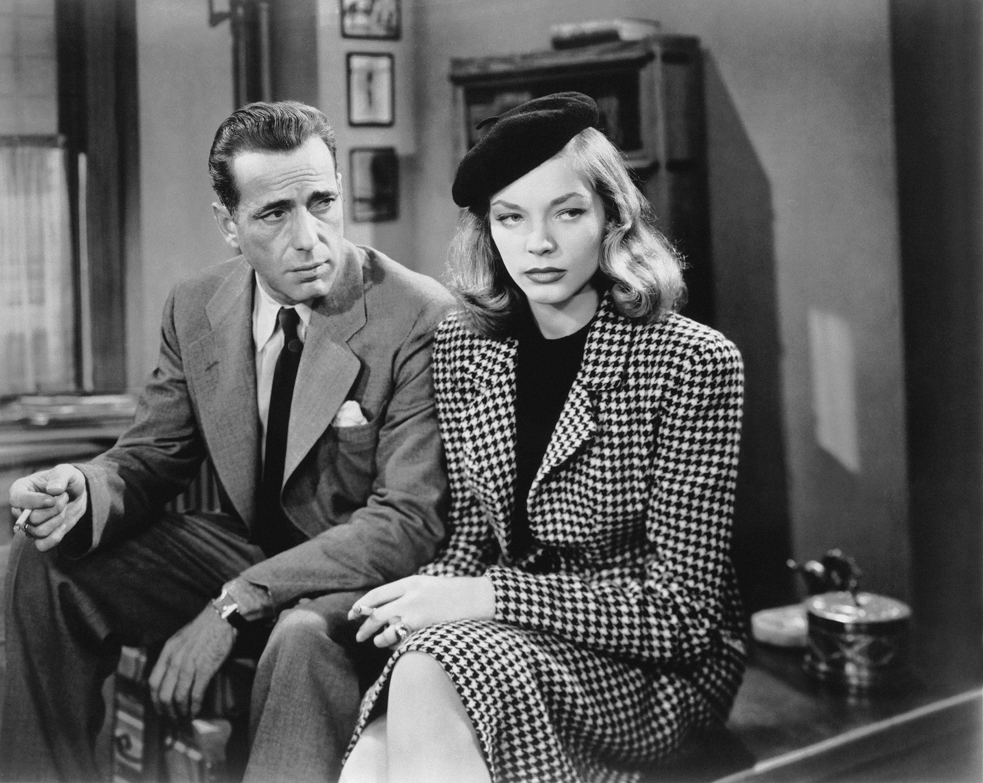 Medium shot from the 1946 movie "The Big Sleep," of Humphrey Bogart as Phillip Marlowe and Lauren Bacall as Vivian Sternwood Rutledge, wearing hat/beret, both sitting on edge of desk and holding cigarette.