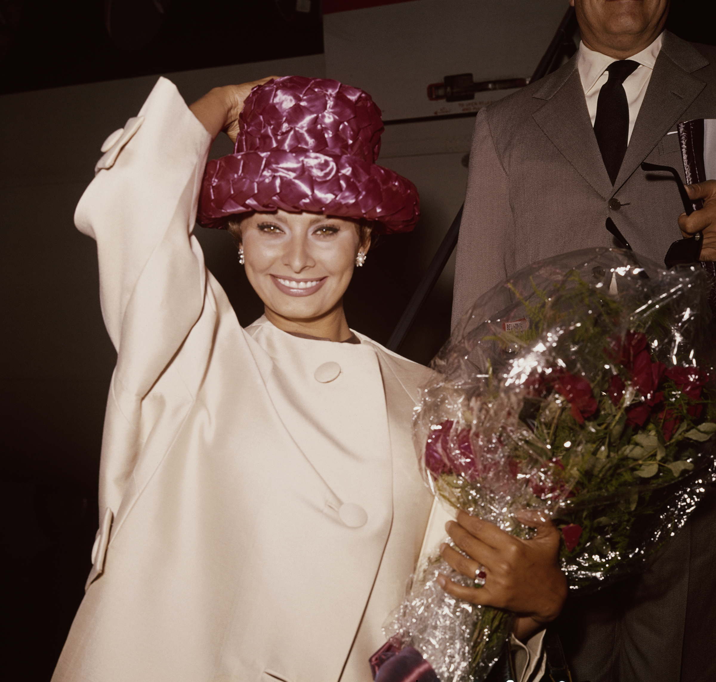 Sophia Loren, Italian actress, holding onto her hat while carrying a bouquet of flowers on her arrival at London Airport, London, England, Great Britain, 15 September 1961.