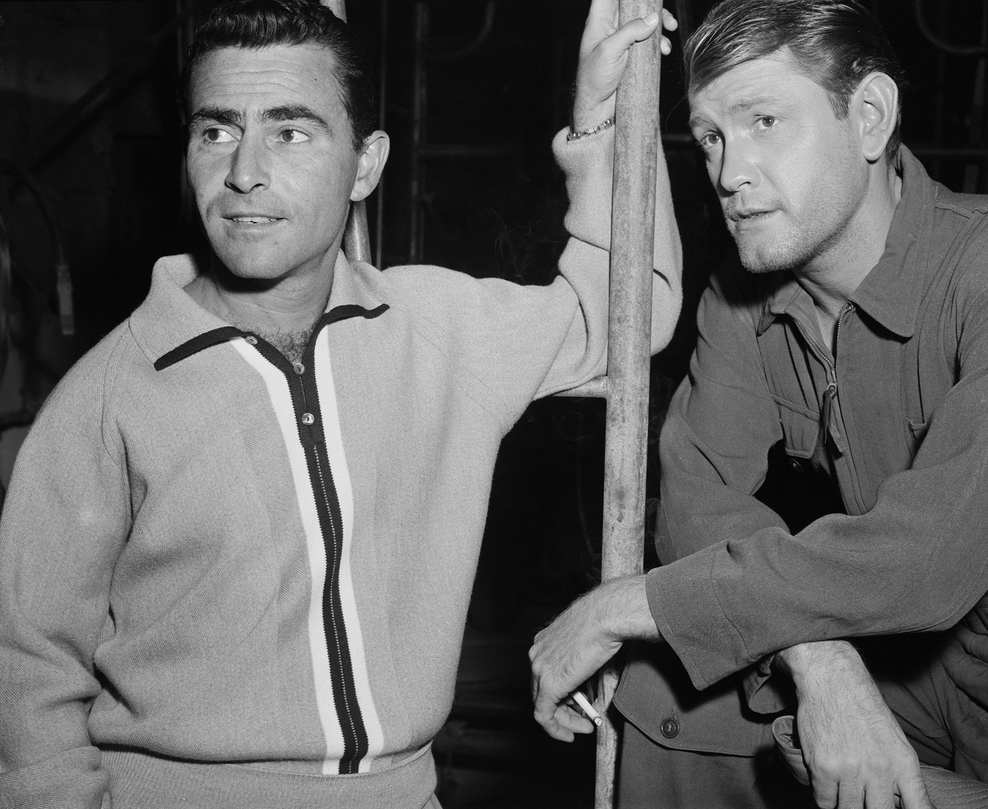 a Dec. 5, 1958, behind the scenes image from between takes on the set of the first "Twilight Zone" episode, "Where Is Everybody?" It is a medium shot of series creator Rod Serling, standing on left, wearing a zip-up white sweater and leaning against a pole with his right arm up against it, and episode star Earl Holliman on the right, with his right leg up on some support and leaning forward and resting his arms on the right knee.