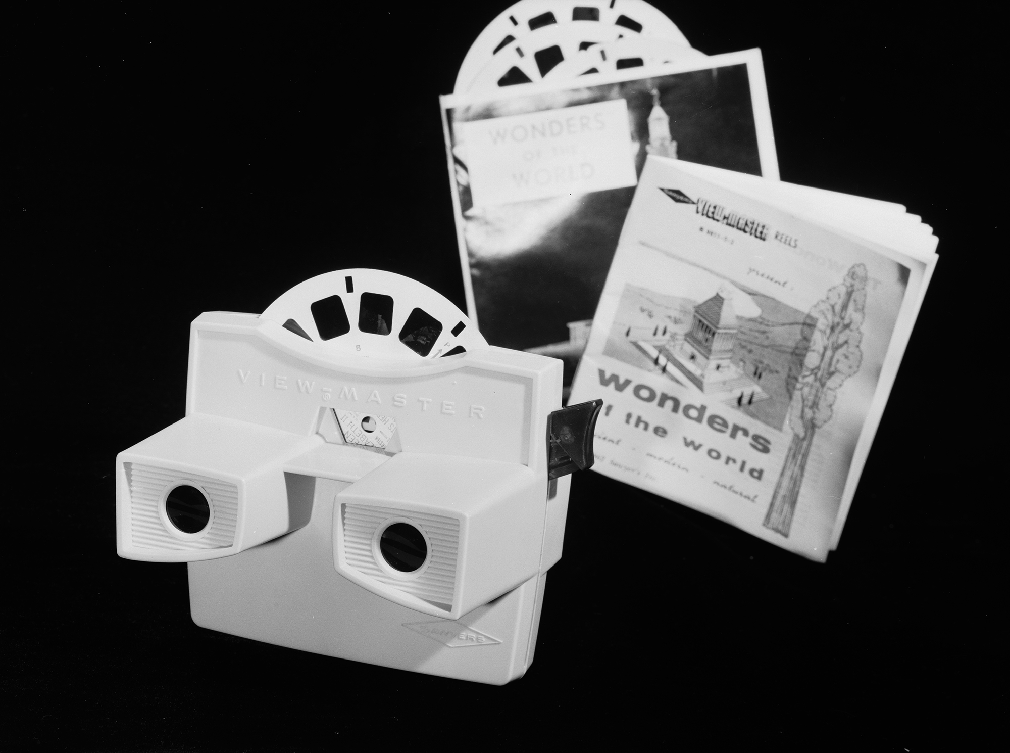 black and white 1963 image of a View-Master camera set against a black backdrop. The camera/projector is on the left, with viewfinder facing outward and an image reel peeking out of it. just behind it is the package, booklet and other reels for "Wonders of the World."
