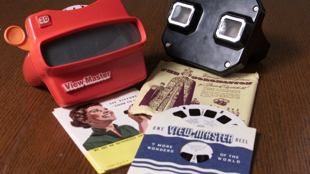 2001 image of a fairly modern, red plastic View-Master camera (on upper left, face down on a table), next to an old, black bakelite View-Master camera (face down just to the right of the red one). Below each camera are three View-Master image reels in jackets, with one reel slightly peeking out of its jacket to reveal the black image squares on the outside of the white reel.
