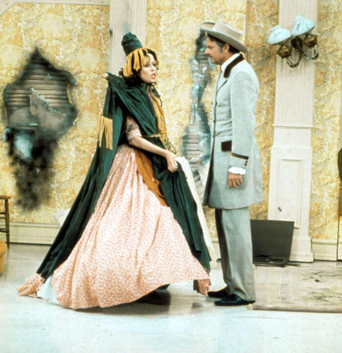 Image from the "Gone with the wind!" Sketch, a parody of "Gone with the wind" from a November 1976 episode "The Carol Burnett Show." On the left is Carol Burnett "Starlett O'Hara," wears a dress made from a drape (complete with rod just behind the neck). Directly in front of her, on the right in the photo, is Harvey Korman "Rat Butler," dressed in a Civil War-era Confederate-style uniform.