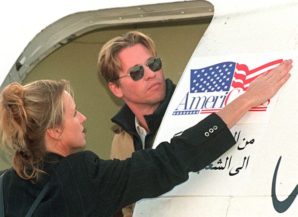 American movie star Val Kilmer (R) watches 28 April at Amman airport as an unidentified woman sticks the logo of US private charity AmeriCares on the door of a Jordanian cargo plane loaded with medical supplies and food donated to Iraq. The 34 tonnes of medical supplies and basic food stuff are the first airlift of US donations to Iraq, which has been hit by more than seven years of UN sanctions
