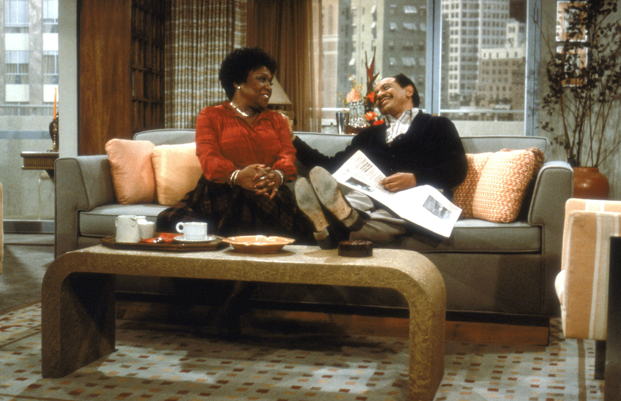 image from the 1970s sitcom "The Jeffersons." It is a long shot of stars Isabel Sanford as "Weezy," sitting on the left on a couch, and Sherman Hemsley as George, sitting on the right. Weezy has her hands clasped over her knee as she looks and smiles at George; George is smiling back, and he has his right hand on Weezy's left shoulder, with a newspaper opened on his lap and his feet up on a coffee table.