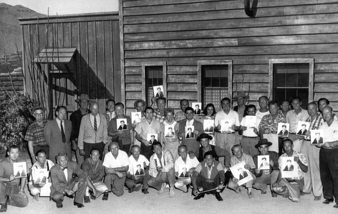 RIO BRAVO, Dean Martin (kneeling seventh from left), director Howard Hawks (standing third from left in sweater), John Wayne (behind Hawks in cowboy hat) with crew members holding pictures of Jerry Lewis on set, 1959