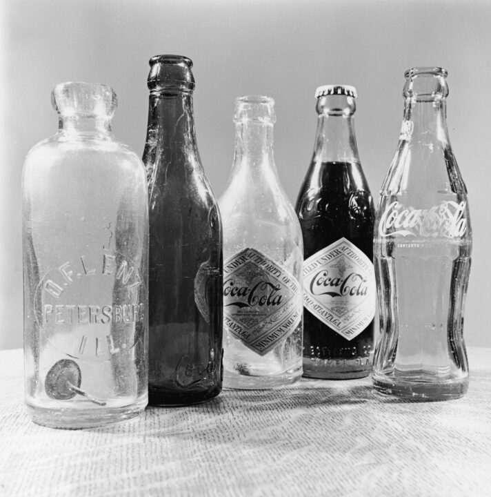 Photograph shows five soda bottles, 1980s. From left, a straight side, center slug bottle labelled 'O.F. Lenz, Peterson, Ill.' dating from the early 1900s (and similar to early Coca-Cola bottles), a straight side dark (possibly amber) bottle with Coca-Cola in script on raised letters at the bottom of the glass dating from approximately 1902 - 1915, a pair of clear, straight side bottles with diamond-shaped paper Coca-Cola logos used in the late 1910s (the bottle with liquid in it additionally features Coca-Cola's raised glass script letters), and a contour shaped Coca-Cola bottle which which was first used around 1915 (though this one, with the painted logo likely dates from the late 1950s).