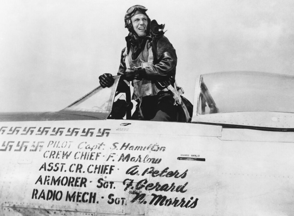 black and white image from the 1948 movie "Fighter Squadron." Star Robert Stack, dressed in a World War II era U.S. Air Force outfit, with bomber jacket and helmet, is standing up in the open cockpit of his fighter plane. Below him, we can see the plane adorned with a number of Nazi swastikas indicating the number of German planes his crew has shot down. Next to those are written out the names of the bomber crew's members and their positions.