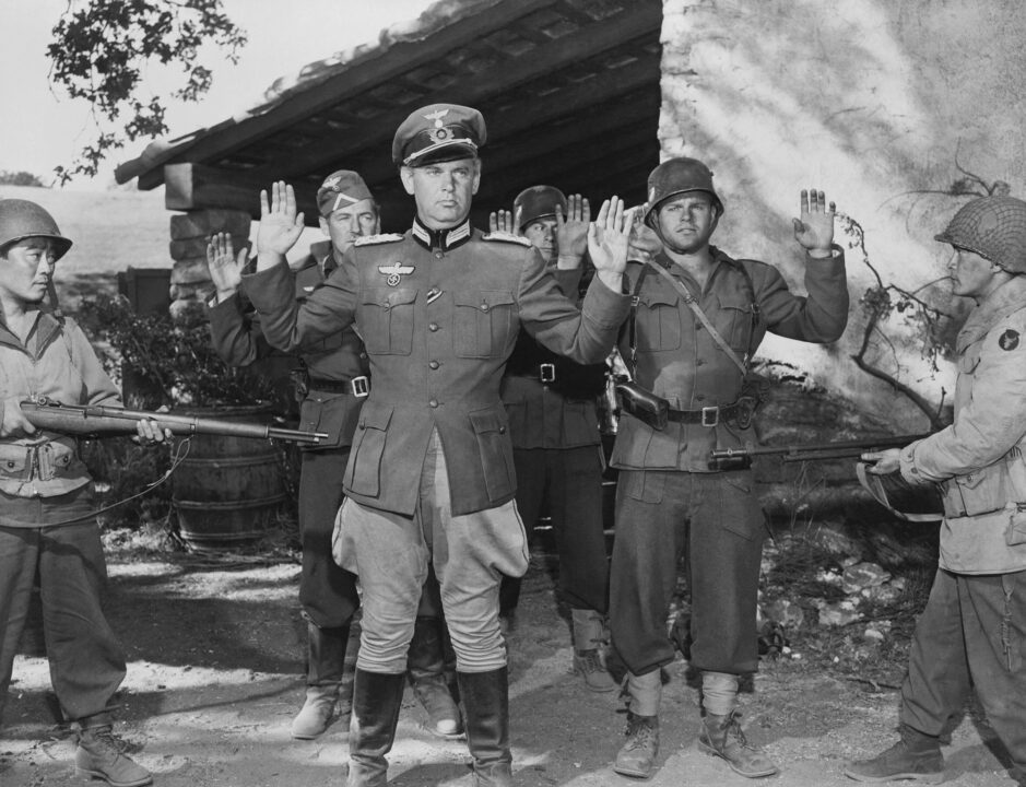 black and white photo from the 1951 movie "Go for Broke!" The film is about the real-life 442nd Regimental Combat Team of World War II, made up of Nisei soldiers. In the photo, real-life 442nd veterans Ken Okamoto and George Miki are pictured in their film roles as soldiers; Okamoto on the far left and Miki on the far right. They are dressed in World War II soldier uniforms and holding guns pointed at surrendering German officers.