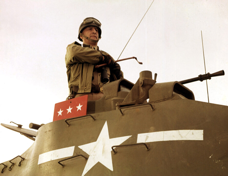image from the 1970 movie "Patton." Pictured is George C. Scott as World War II General George S. Patton, who is standing upright in a tank. He is wearing his general's outfit, and a helmet with goggles up on it. The tank has a white stripe and star across its body.