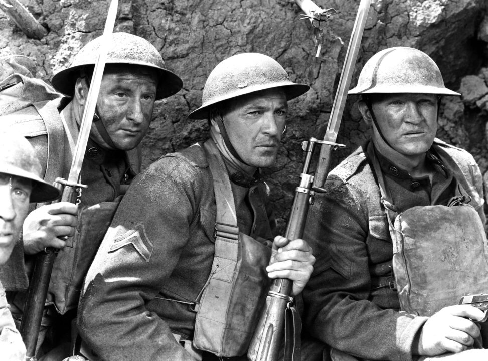 black and white image from the 1941 movie "Sergeant York." From left to right are George Tobias, Gary Cooper (as Sgt. York) and Joe Sawyer as American soldiers crouching in a trench during World War I, holding guns with bayonets and wearing helmets and uniforms of the time.