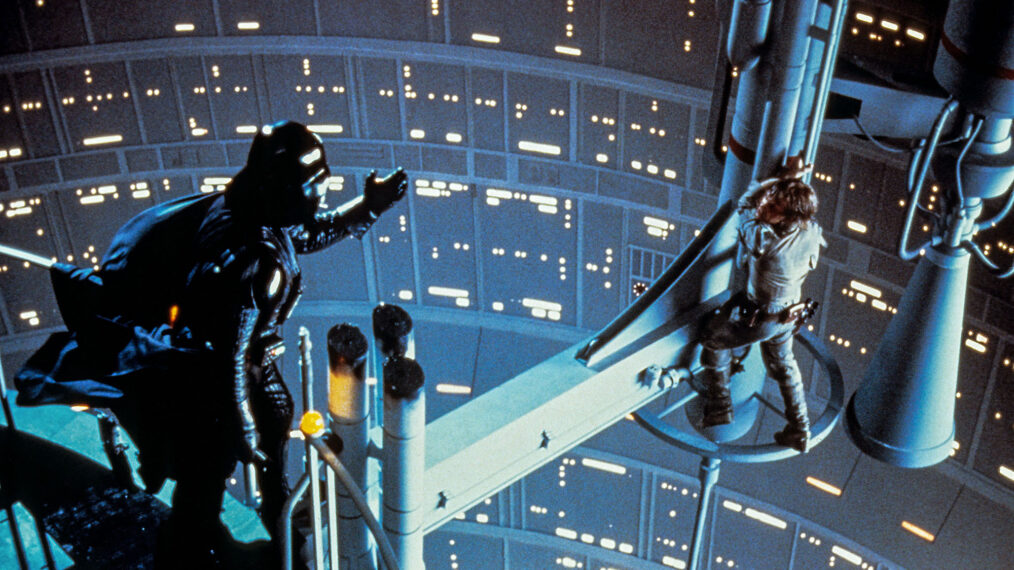 image from the 1980 Star Wars movie The Empire Strikes Back. It is an overhead look at Darth Vader, dressed in black helmet, cape and armor, standing on the left. In his right hand he his holding his lightsaber, which has been turned off. His left arm is reaching out toward Luke Skywalker (Mark Hamill), on the right of the photo. Luke is clinging to the edge of a bridge-like structure, below which is a far drop. He is listening in shock as Vader communicates to him that he is his father.