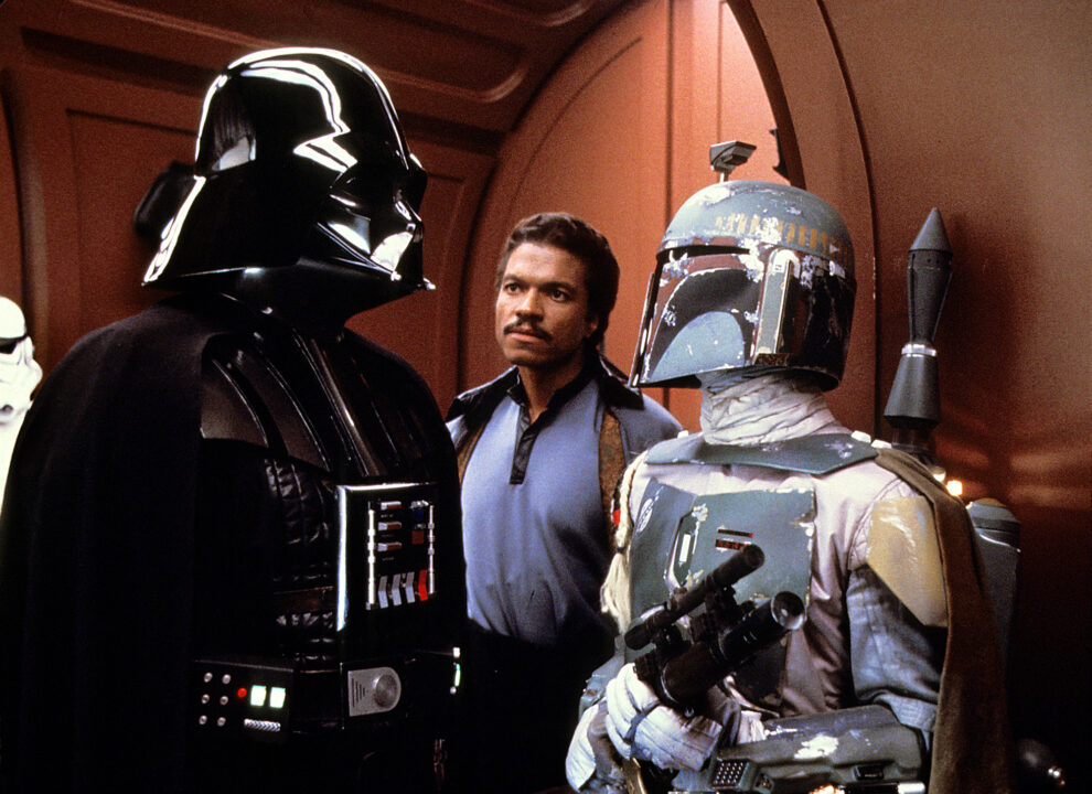 medium shot of a scene from the 1980 Star Wars movie "The Empire Strikes Back." On the left is Darth Vader, dressed in his black cape and helmet; to his left, in the center and a bit back, is Lando Calrissian (Billy Dee Williams), dressed in a blue shirt and cloak, looking with concern at Vader; and to his left, near the front of the photo and looking at and speaking with Vader, is bounty hunter Boba Fett, brandishing a laser blaster in both hands and wearing a banged-up helment and multi-colored (gray/green/yellow/red) armored suit that has seen a lot of wear and tear.