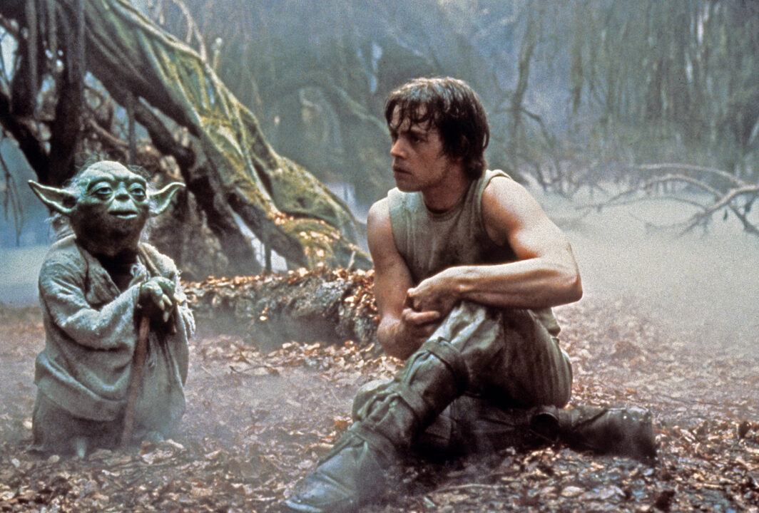 image from the 1980 Star Wars movie The Empire Strikes Back. It depicts a scene on the swampy world of Dagobah, where Jedi master Yoda, a short, green-skinned alien-looking being wearing a light tan robe, is standing on the left, instructing a sweaty and tired-looking Luke Skywalker (Mark Hamill), who is seated on the right of the photo, looking somewhat dejected.