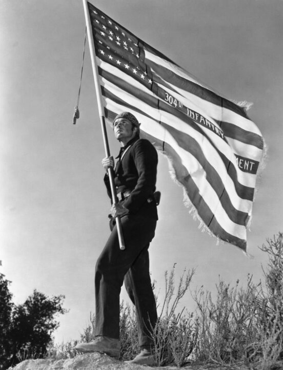 black and white image from the 1951 movie "The Red Badge of Courage." It is a vertical shot of star Audie Murphy, in character as a Civil War Union soldier, holding an American flag in both hands.