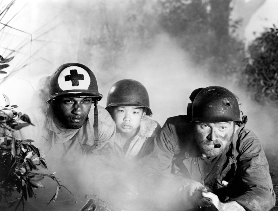 black and white image from the 1951 movie "The Steel Helmet." From left to right, crouching down and looking intently ahead at something during a Korean War battle are, left to right, characters played by James Edwards, a Black man wearing a helmet adorned with a cross indicating he is medical personnel; in the center, a Korean boy, also wearing a helmet; and on the left, a character played by Gene Evans, wearing a helmet and with a cigar in his mouth. Evans has a rifle in his hands.