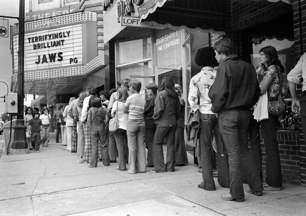 View of a line of moviegoers outside the State Theatre (at 233 South State Street), Ann Arbor, Michigan, July 14, 1975. The cinema marquee advertises the film 'Jaws.'
