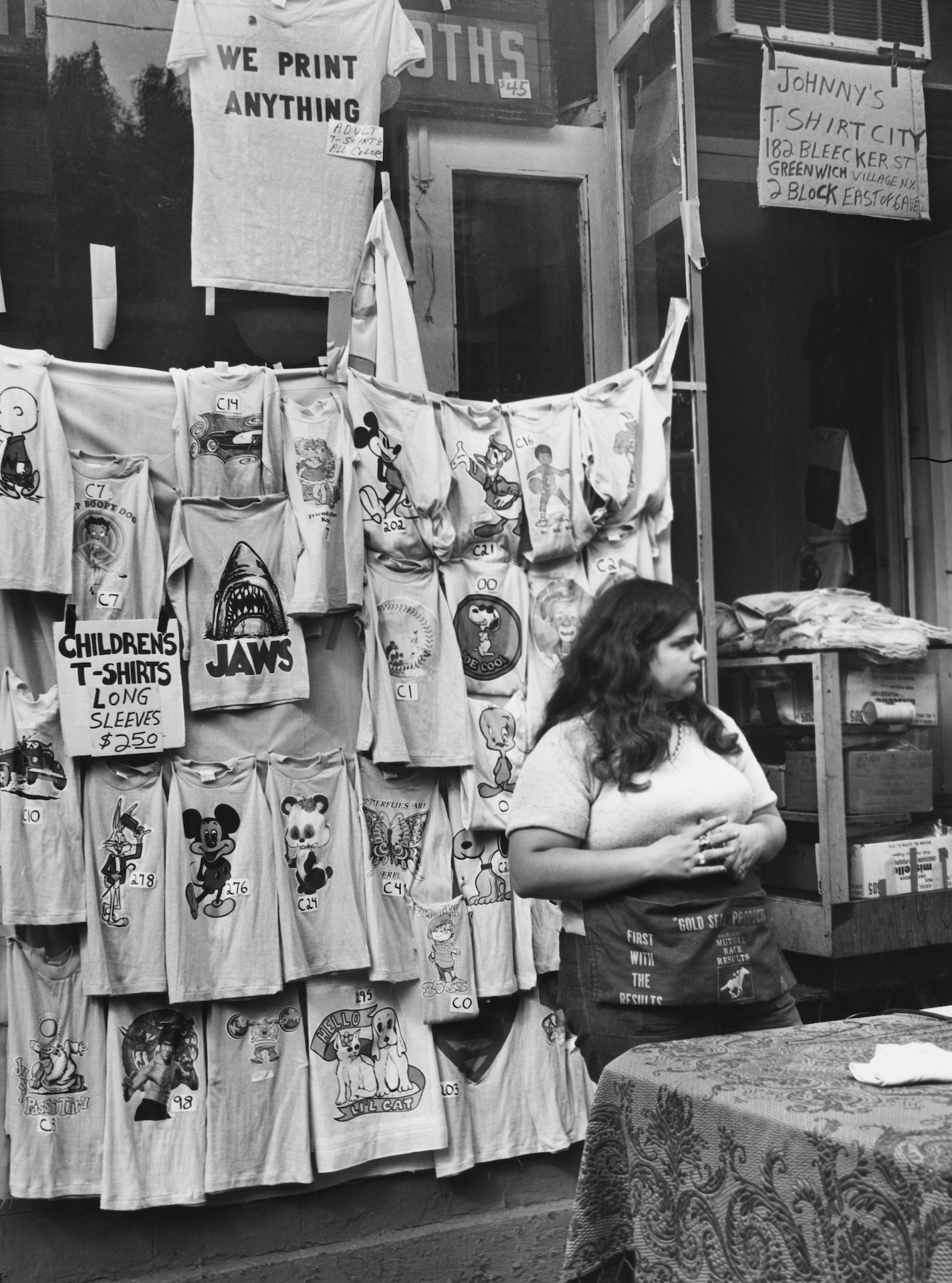 T-shirts featuring images of Jaws, Snoopy, Mickey Mouse, Bugs Bunny, Donald Duck, and Tweety displayed for sale at Johnny's T-Shirt City, on Bleecker Street in the Greenwich Village neighbourhood of Lower Manhattan in New York City, New York, October 1975. Signs read 'We Print Anything', 'Children's T-Shirts Long Sleeves $2 50', and 'Johnny's T-Shirt City, 182 Bleecker Street, Greenwich Village, 2 Block East of 6 Ave'.