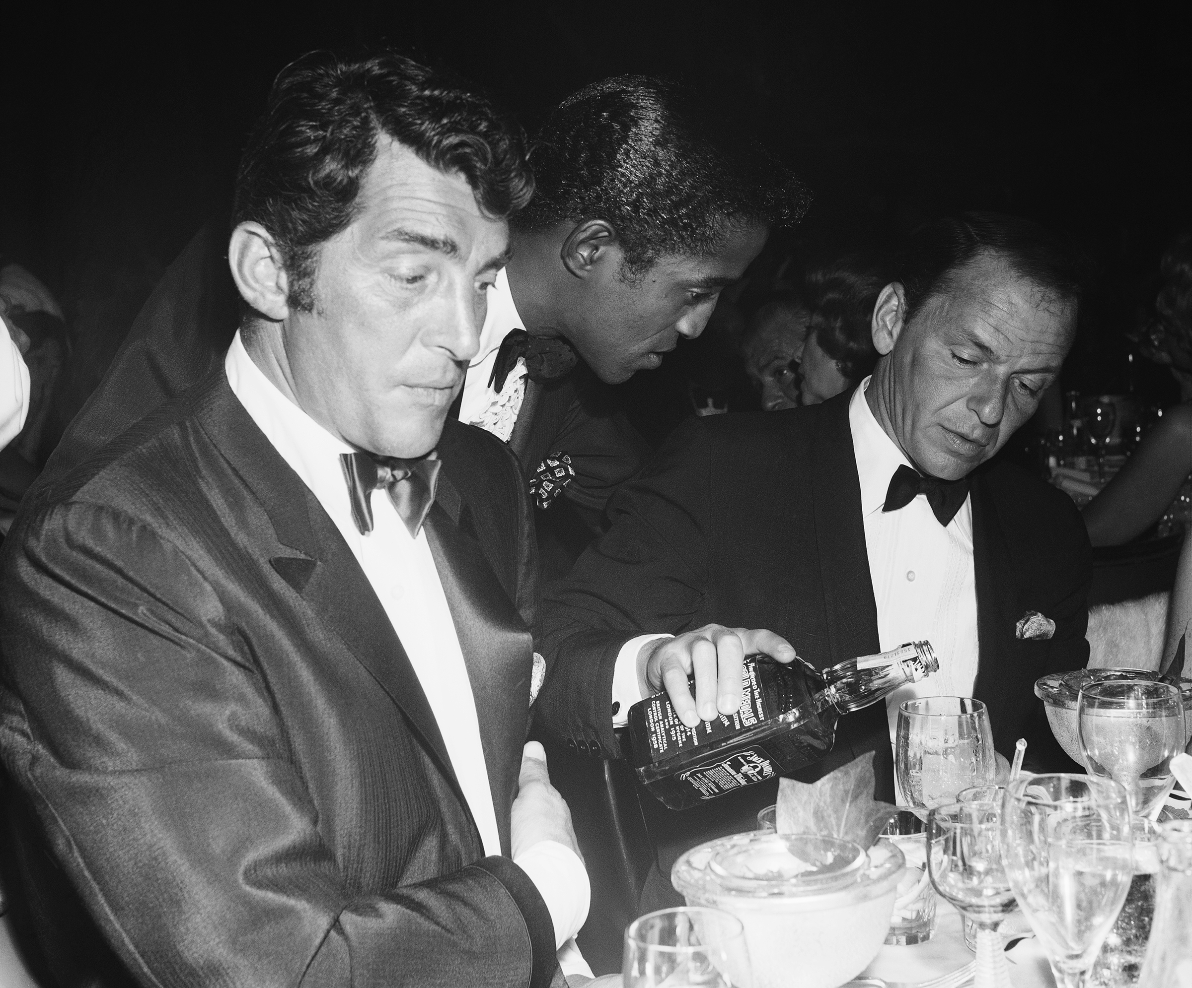 Singer Dean Martin and Sammy Davis, Jr. sit at a table as fellow singer and Rat Pack member Frank Sinatra pours Jack Daniels from a bottle at the Cocoanut Grove during Eddie Fisher's opening night on July 25, 1961 in Los Angeles, California.
