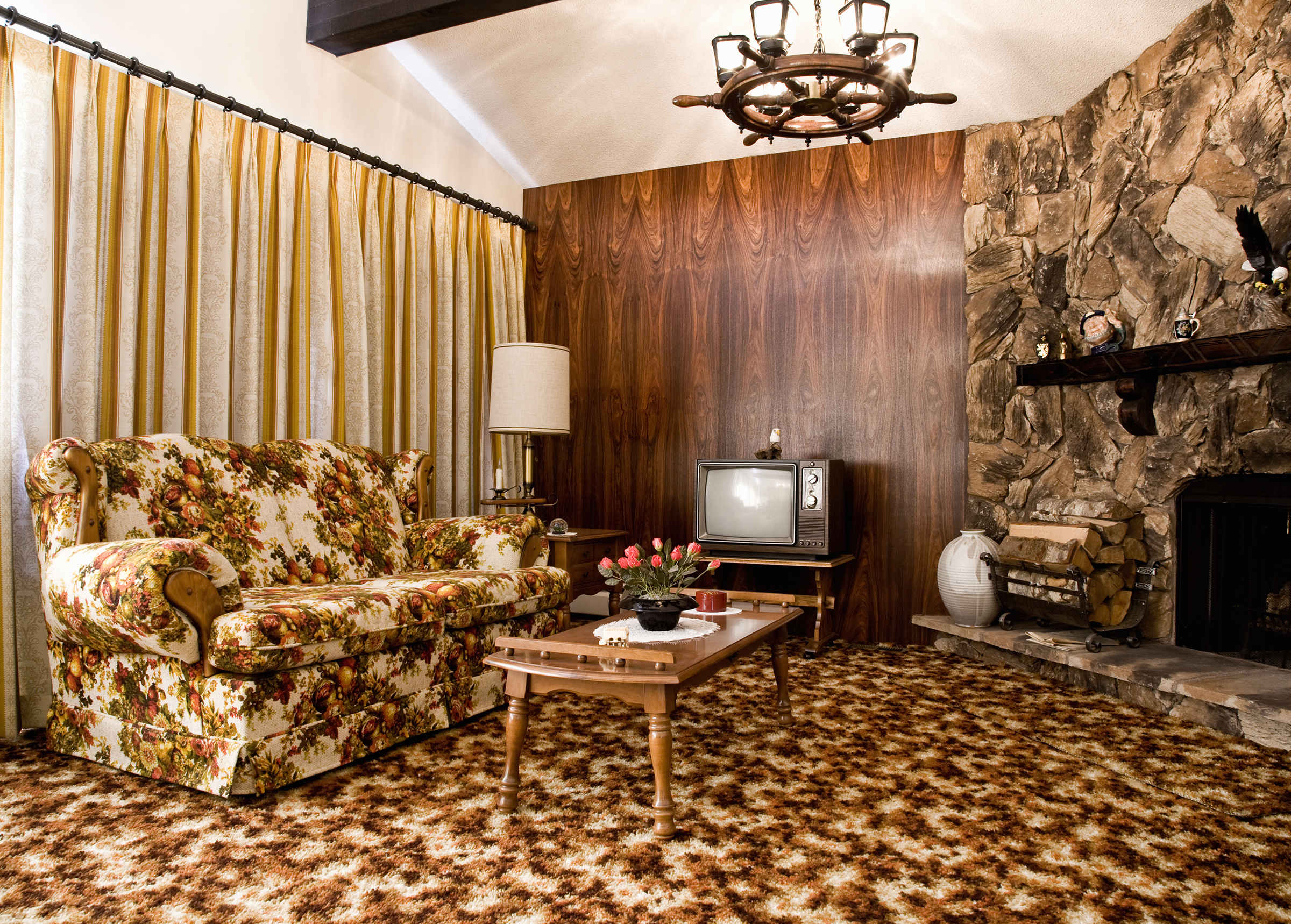 1970s era living room - stock photo A wide, horizontal shot of a retro living room with a centered composition, showcasing a floral-patterned sofa and armchair, a wooden coffee table with a potted plant, and a stone fireplace with a television set against a wood-paneled wall, all under warm, soft lighting.